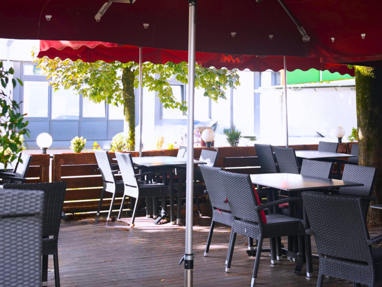 Großer, modern möblierter Außenbereich des Restaurants mit dunklen Tischen, Stühlen und roten Sonnenschirmen.